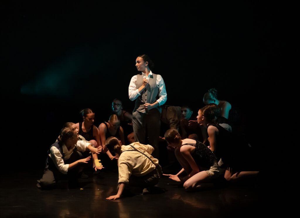 One dancers stood in the middle holding their tie and the bottom of the waistcoat. The other dancers form a circle around her, looking up at her.