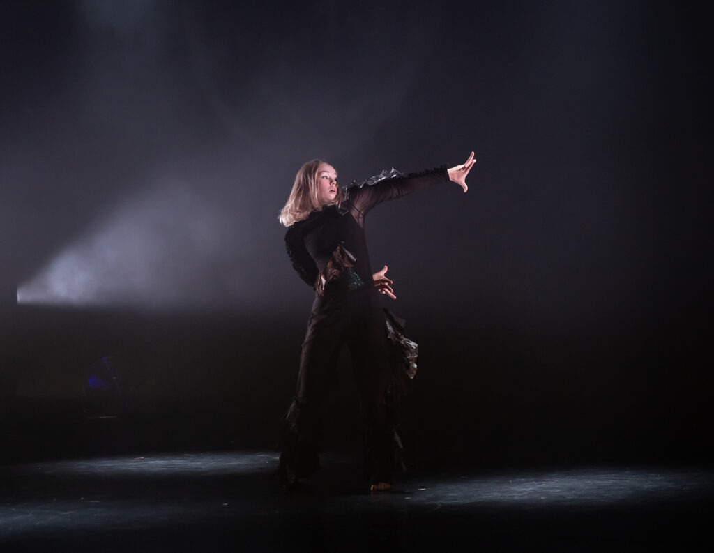 A dancer on stage in all black. Hands are swung out to the right, one hand behind her back.