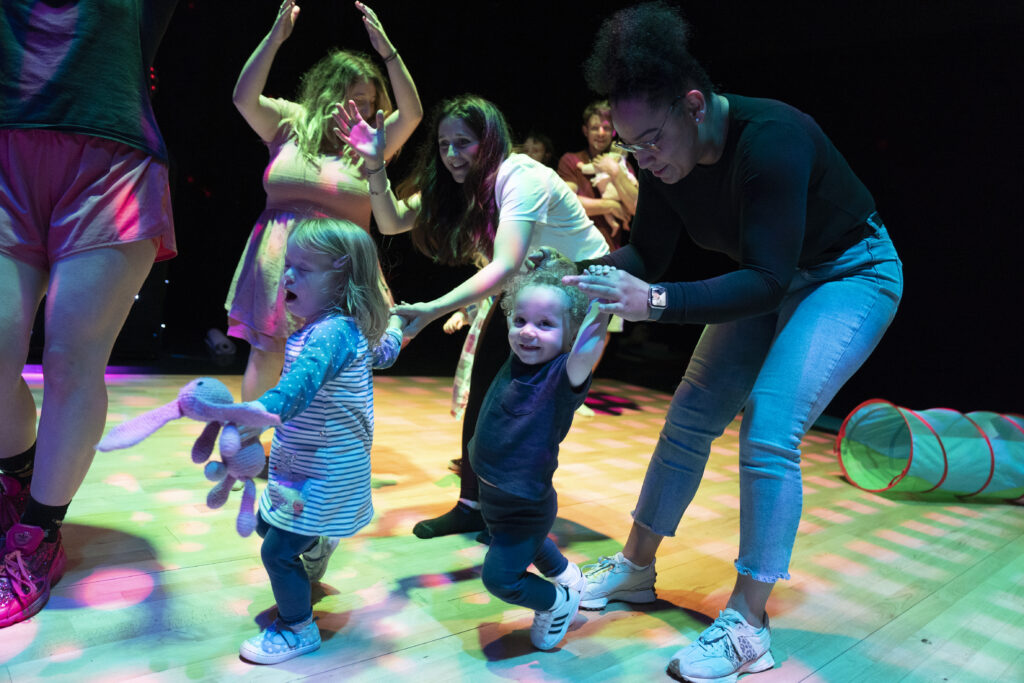 Two adults dancing with two children. One child is moving towards the camera. One child is holding their adults hands above their head.