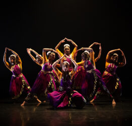 A group of dancers from Bhadram School of Dance together in the middle of a dark stage. Their costumes are rich purple and yellow in colour.
