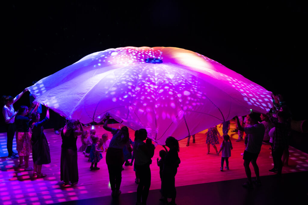 A group of people in a dark theatre. Lights highlight the parachute. Adults are stood around holding the parachute with young people running underneath.